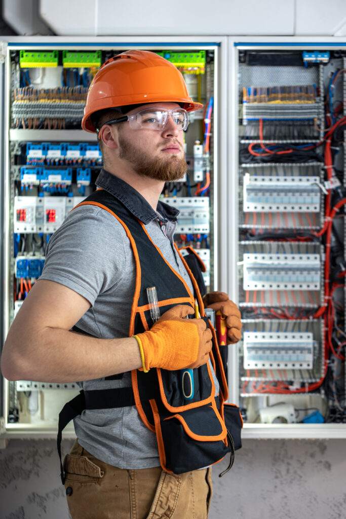 Electrician man in overalls works in the switchboard. Portrait of an electrician in overalls. High quality photo. Safety glasses, hard hat and electrical shield.